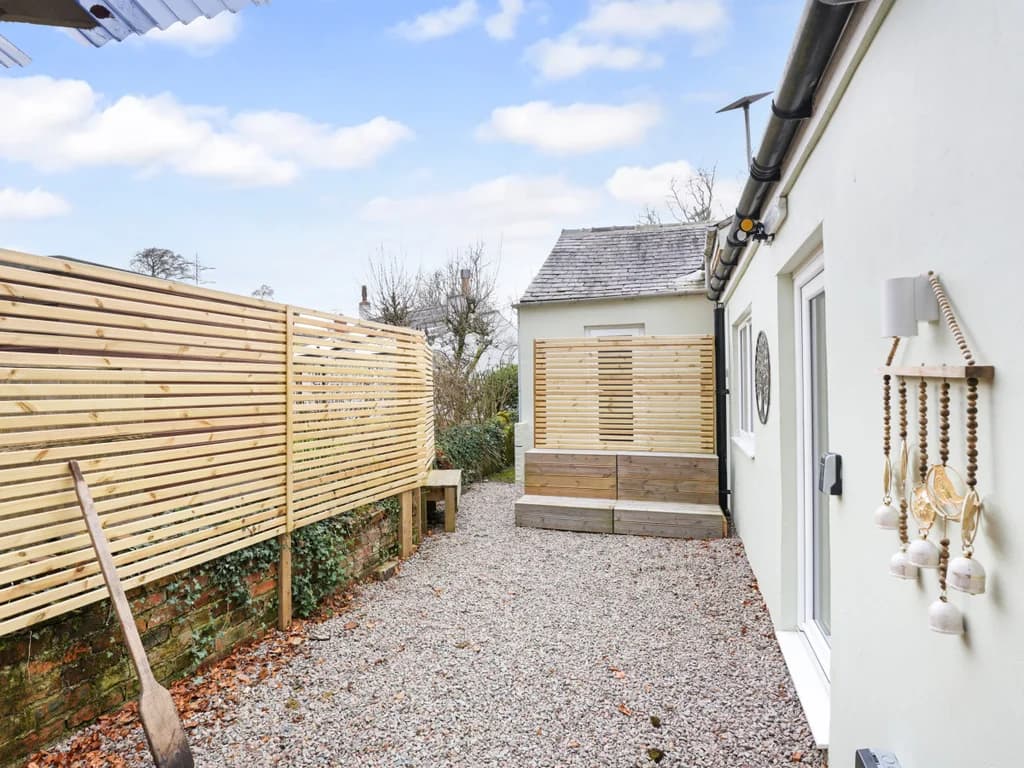 A private gravel courtyard features a slatted wooden privacy fence, built-in timber seating, and decorative wind chimes hanging against the white exterior wall of the cottage.