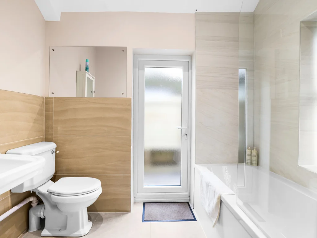 This modern bathroom features a sleek white bathtub with a glass screen, neutral-toned wall tiling, and a frosted glass door that leads to the outdoors.