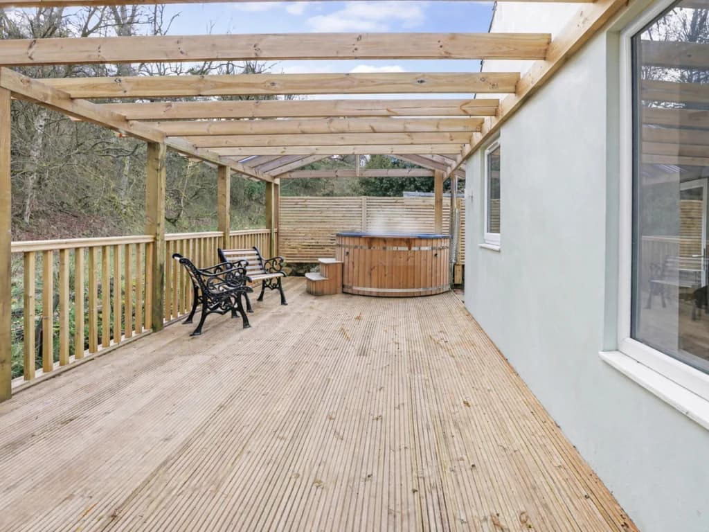 A steaming wood-fired hot tub sits on a wooden deck under a transparent-roofed gazebo, offering a cozy outdoor retreat surrounded by a natural, wooded landscape.