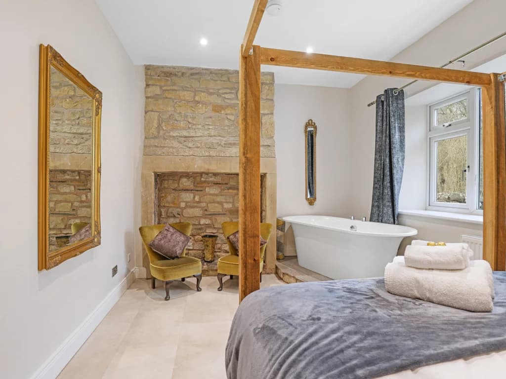 This elegant bedroom blends rustic stone features with modern luxury, highlighting a wooden four-poster bed and a freestanding bathtub positioned by a bright window.