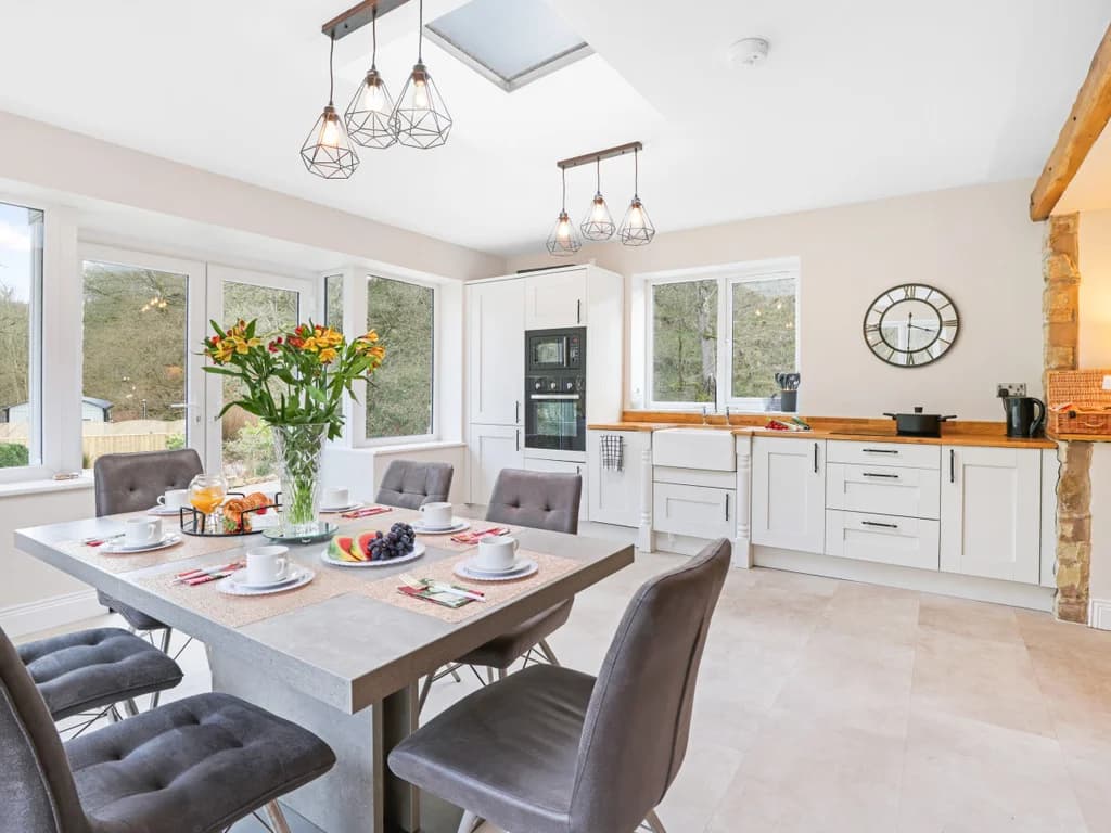 This bright, contemporary kitchen and dining area features white cabinetry, a skylight, and a large dining table set for breakfast, all illuminated by stylish geometric pendant lights.