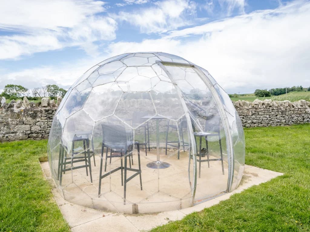 Transparent garden dining dome set on lawn beside stone walls