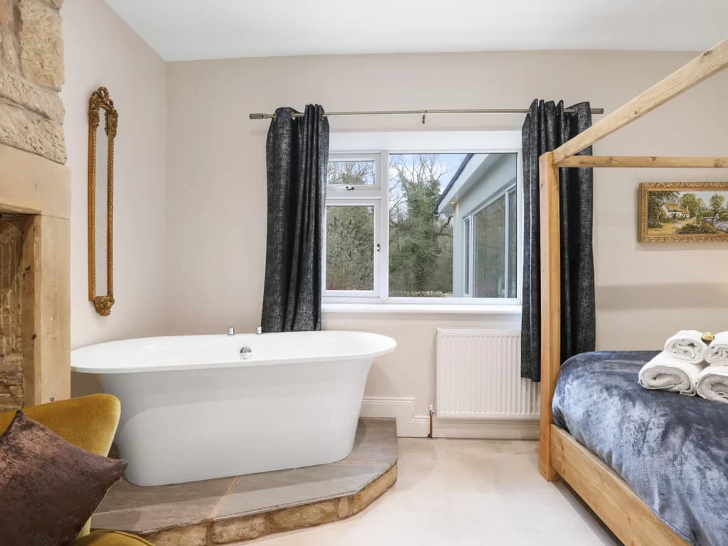 A freestanding soaking tub sits on a raised stone plinth in this elegant bedroom, positioned between a rustic fireplace and a wooden four-poster bed.