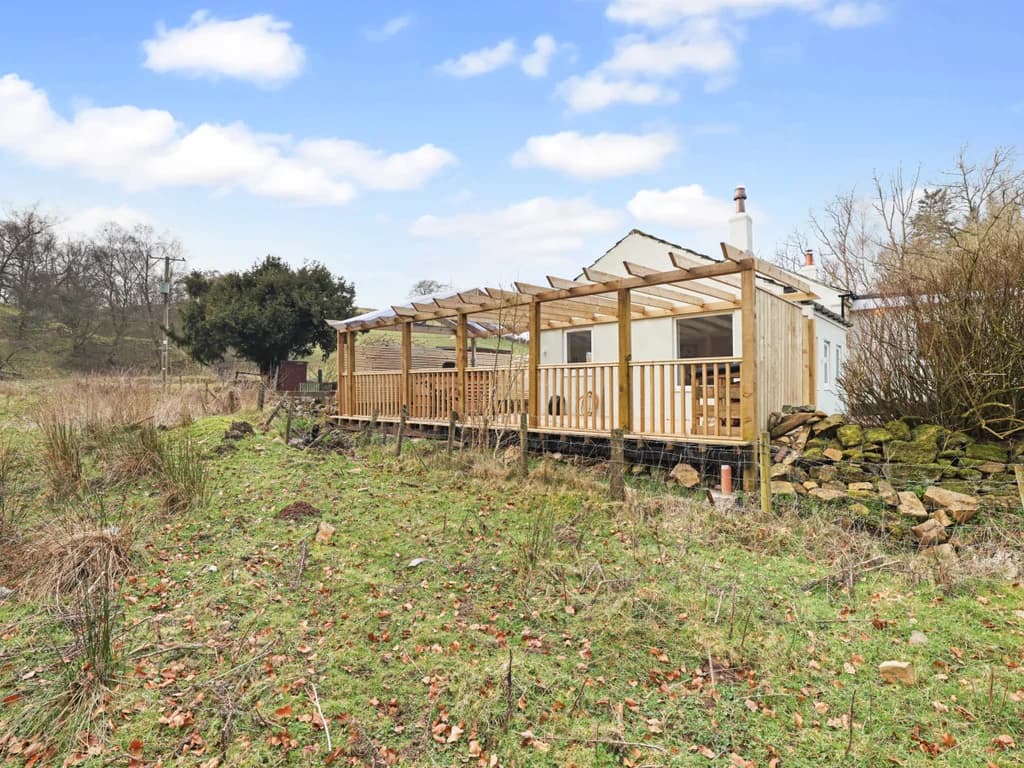 A spacious wooden deck with a pergola-style roof extends from the side of this light green cottage, offering a scenic view of the surrounding rural landscape.