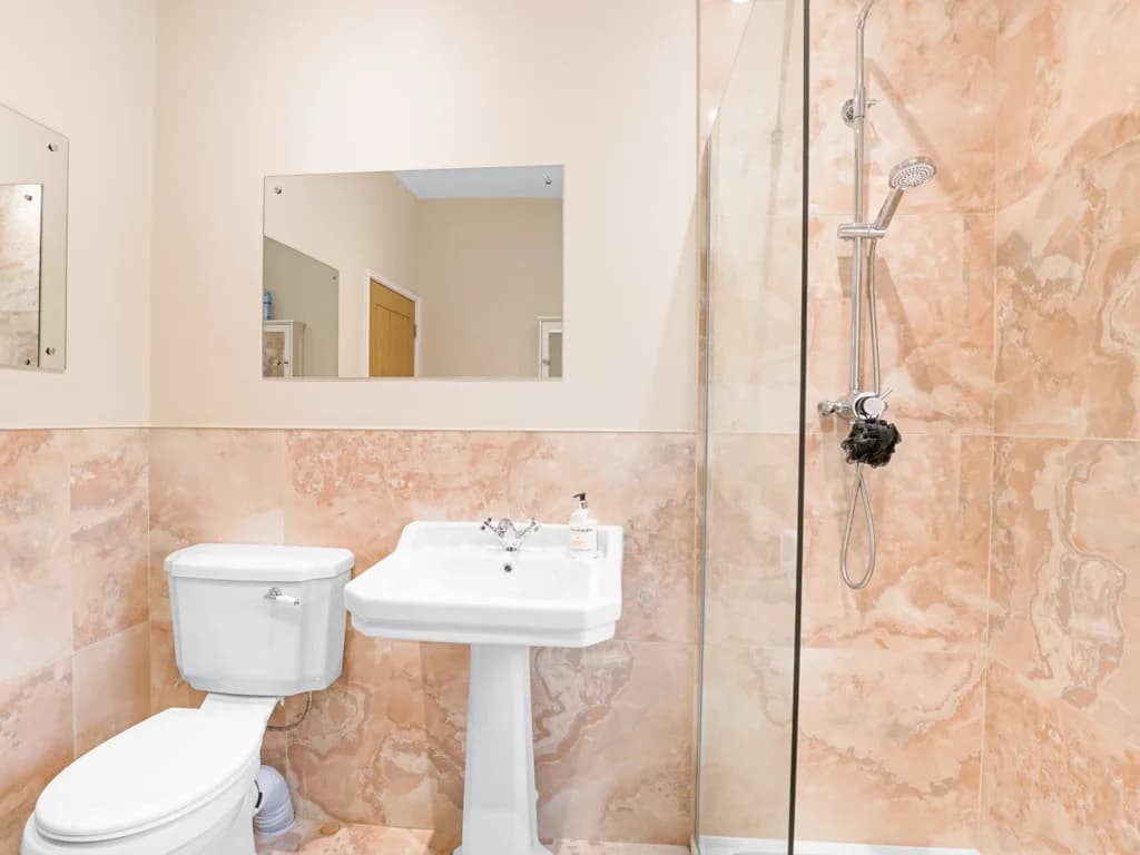 This elegant bathroom features peach-toned marble tiling, a classic pedestal sink, and a modern glass-enclosed shower.
