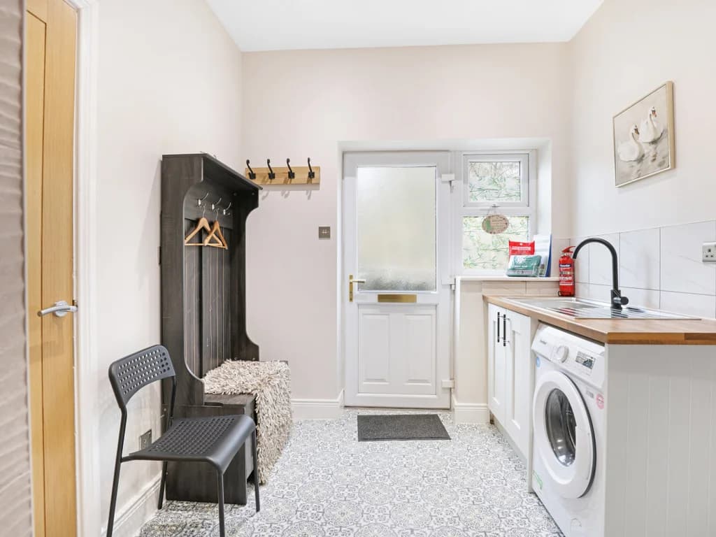 This functional utility room and entryway features patterned tile flooring, a washing machine under a wooden countertop, and a dark wood hall tree with a built-in bench.