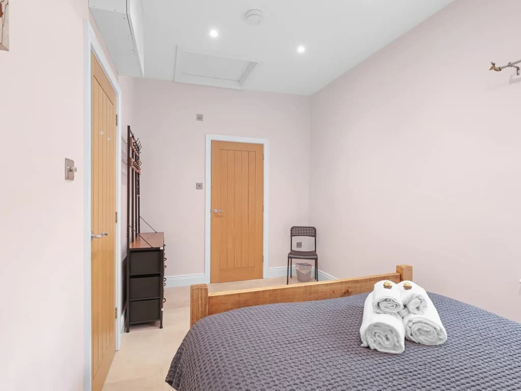 This bedroom features soft pink walls, a wooden bed frame with a textured grey throw, and a modern black valet stand for storage.