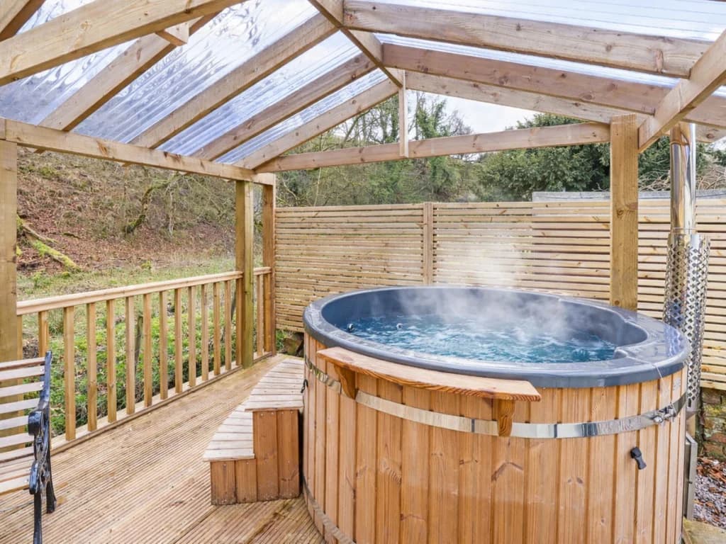 A steaming wood-fired hot tub sits on a wooden deck under a transparent-roofed gazebo, offering a cozy outdoor retreat surrounded by a natural, wooded landscape.