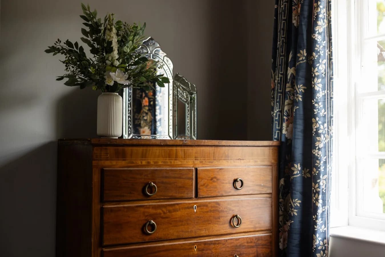 Walnut chest of drawers with ornate mirror and floral arrangement.