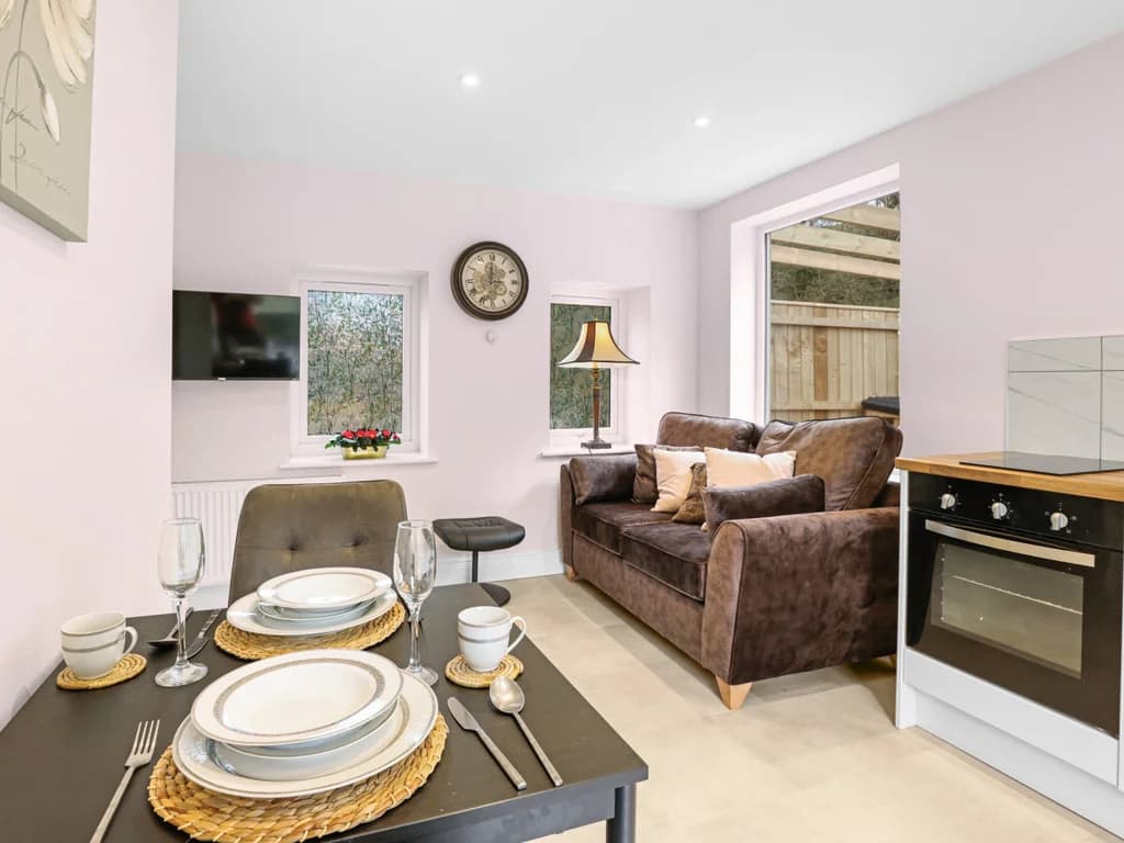 This cosy, open-plan living area features a modern kitchenette, a small dining table set for two, and a plush brown sofa.