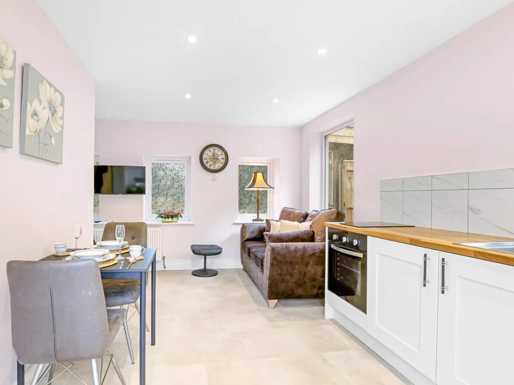 This compact open-plan living area combines a modern white kitchenette with a cosy dining nook and a brown leather sofa set against soft walls.