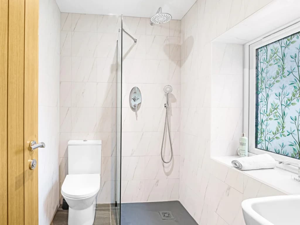 This modern, bright bathroom features a walk-in shower with a glass screen, sleek white tiling, and a window with a decorative leaf-patterned privacy film.