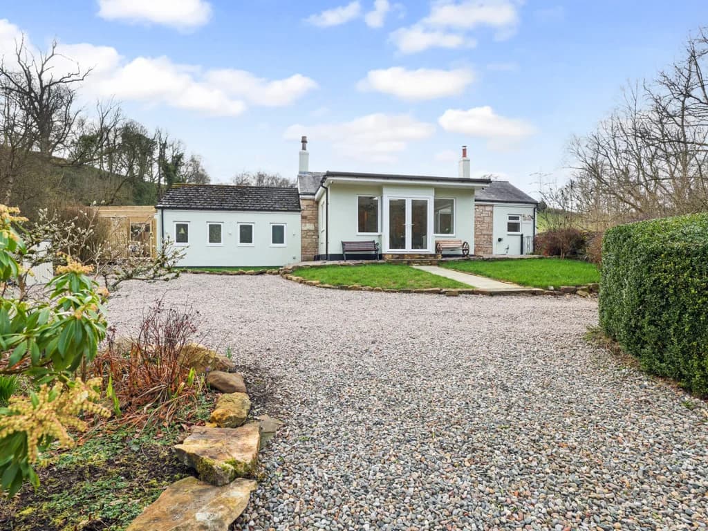 A charming cottage with a modern white extension stands nestled against a wooded hillside, with grass and a gravelled parking area.