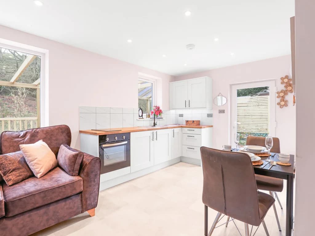 This compact open-plan living area combines a modern white kitchenette with a cosy dining nook and a brown leather sofa.