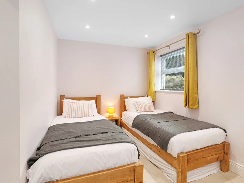 This cosy twin bedroom features two rustic wooden beds with grey throws, accented by vibrant yellow curtains and a warm bedside lamp.