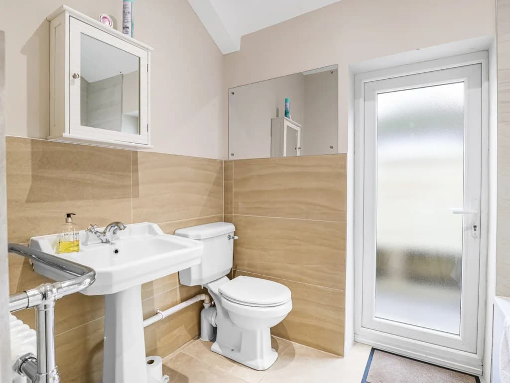 This modern bathroom features a sleek white bathtub with a glass screen, neutral-toned wall tiling, and a frosted glass door that leads to the outdoors.