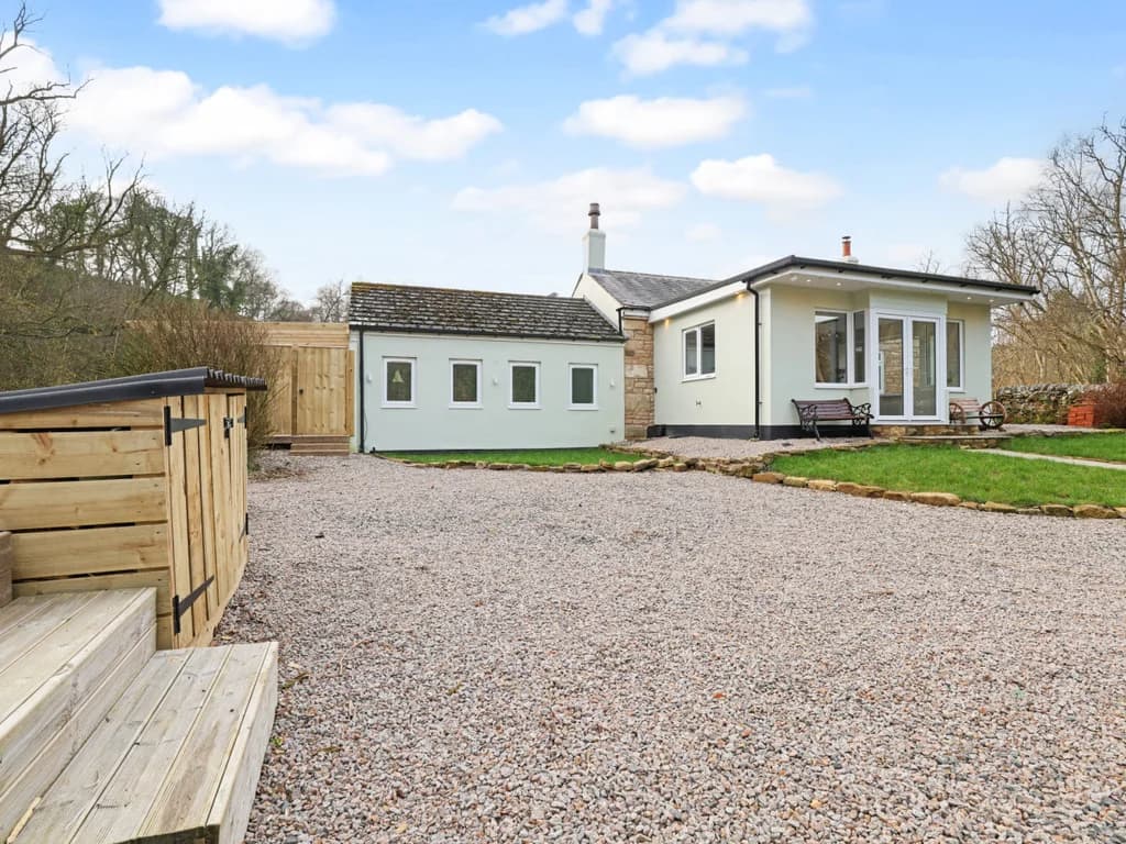 A charming cottage with a modern white extension stands nestled against a wooded hillside, with grass and a gravelled parking area.