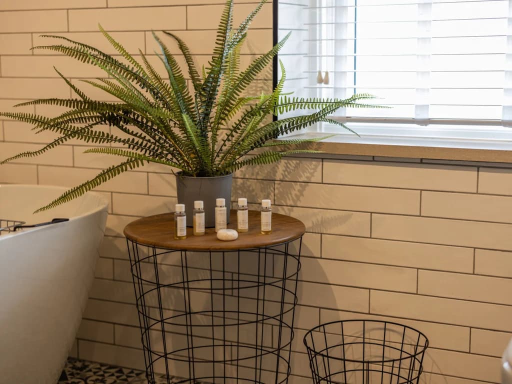 A potted fern and a selection of travel-sized toiletries sit on a wire-frame side table in a bright, modern bathroom featuring white subway tiles and a freestanding tub.