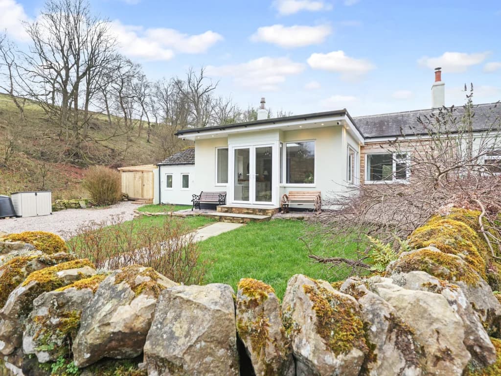 A charming cottage with a modern white extension stands nestled against a wooded hillside, framed by a rustic stone wall and a green lawn.