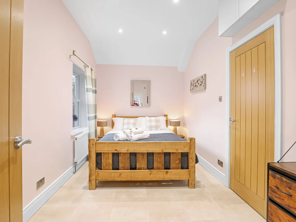 Soft pink walls and a warm wooden bed frame create an inviting atmosphere in this cosy bedroom, complete with plaid pillows and soft bedside lighting.
