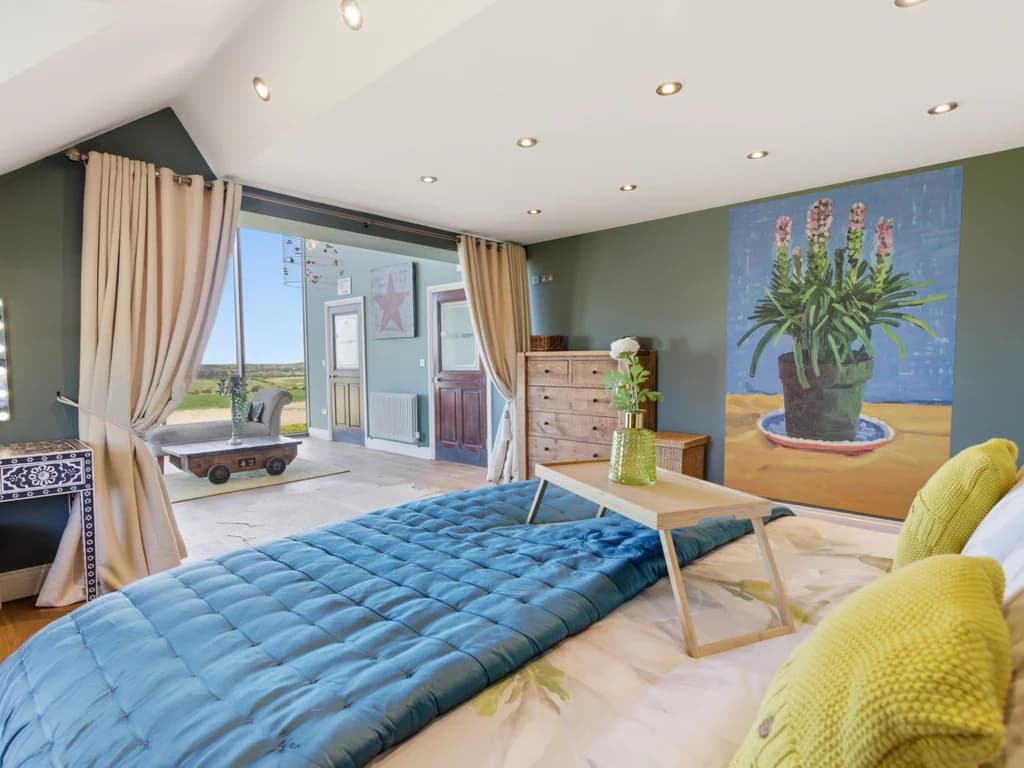 This eclectic bedroom features a blue quilted bedspread and a wooden breakfast tray, set against sage green walls adorned with a large floral painting and an open view into a sunlit lounge area.
