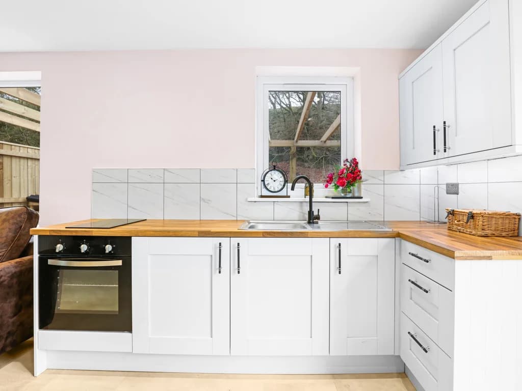 This bright, modern kitchen features white cabinetry and wood countertops, with a large window and a glass door that looks out onto a private outdoor area.